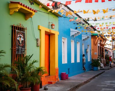 Colorful street scene with vibrant green, orange, and blue buildings, festive hanging flags, and people strolling, conveying a lively, cheerful atmosphere.