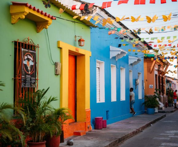 Colorful street scene with vibrant green, orange, and blue buildings, festive hanging flags, and people strolling, conveying a lively, cheerful atmosphere.