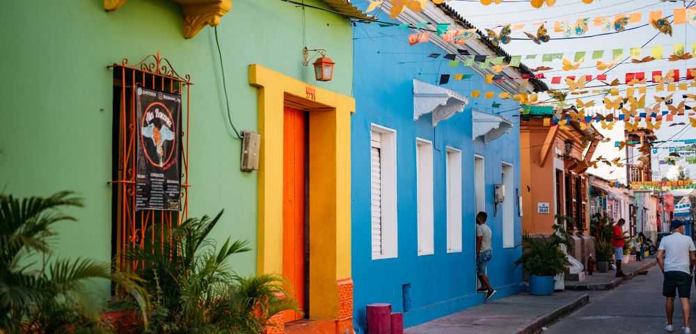 Colorful street scene with vibrant green, orange, and blue buildings, festive hanging flags, and people strolling, conveying a lively, cheerful atmosphere.