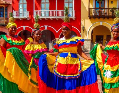 Four women in vivid traditional dresses stand joyfully in front of colorful colonial buildings. Each wears a pineapple headpiece, exuding festive energy.