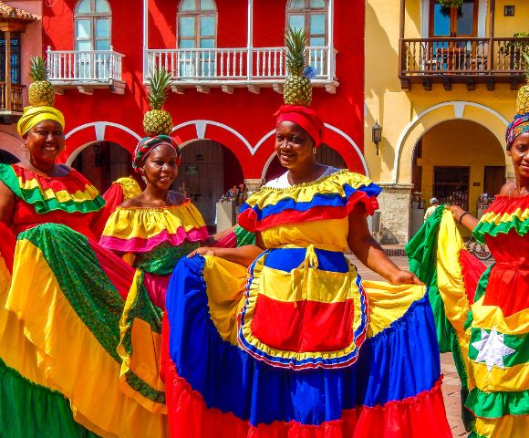 Four women in vivid traditional dresses stand joyfully in front of colorful colonial buildings. Each wears a pineapple headpiece, exuding festive energy.
