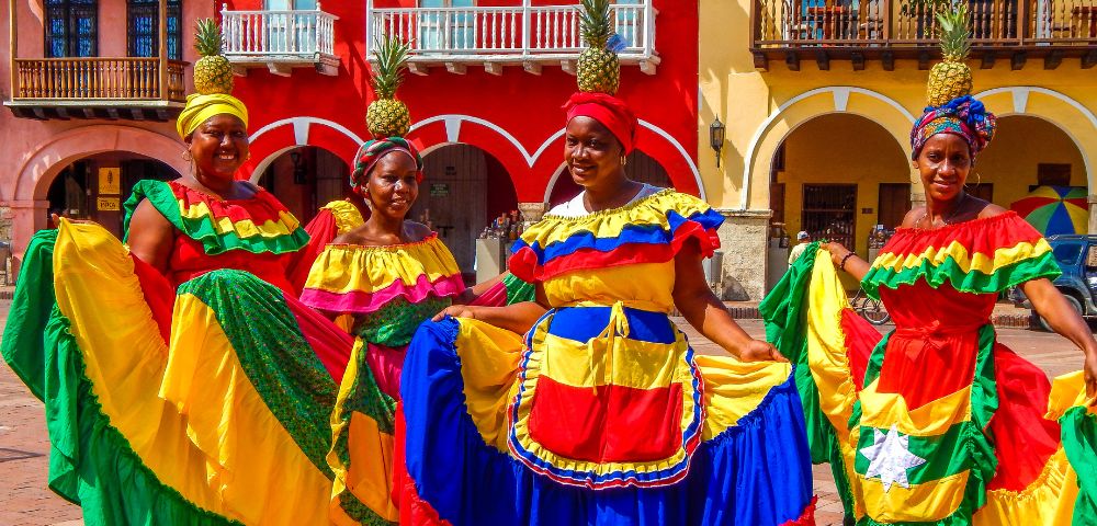 Four women in vivid traditional dresses stand joyfully in front of colorful colonial buildings. Each wears a pineapple headpiece, exuding festive energy.