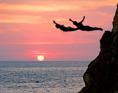 Two people dive from a cliff into the ocean at sunset, silhouetted against a vibrant sky with a glowing sun, conveying adventure and freedom.