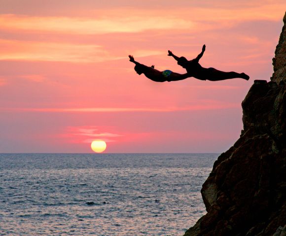 Two people dive from a cliff into the ocean at sunset, silhouetted against a vibrant sky with a glowing sun, conveying adventure and freedom.