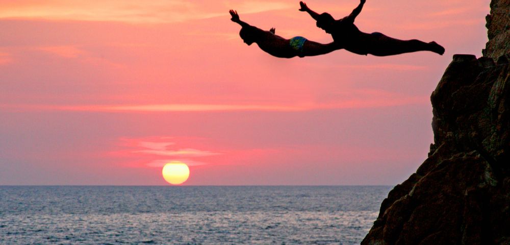 Two people dive from a cliff into the ocean at sunset, silhouetted against a vibrant sky with a glowing sun, conveying adventure and freedom.
