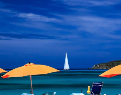 A serene beach scene with vibrant orange umbrellas, turquoise water, and a distant white sailboat under a deep blue sky, evoking a sense of relaxation.