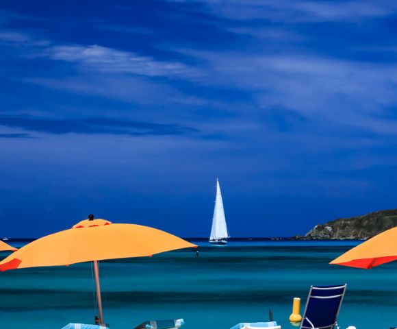 A serene beach scene with vibrant orange umbrellas, turquoise water, and a distant white sailboat under a deep blue sky, evoking a sense of relaxation.