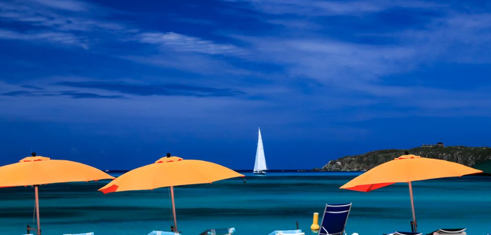 A serene beach scene with vibrant orange umbrellas, turquoise water, and a distant white sailboat under a deep blue sky, evoking a sense of relaxation.