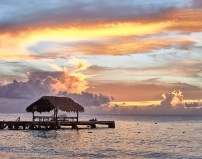 Tropical sunset over calm ocean, with a rustic wooden pier and thatched-roof hut in silhouette. The sky is vibrant with orange, pink, and blue hues.