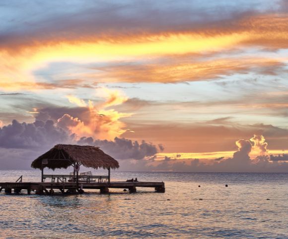 Tropical sunset over calm ocean, with a rustic wooden pier and thatched-roof hut in silhouette. The sky is vibrant with orange, pink, and blue hues.