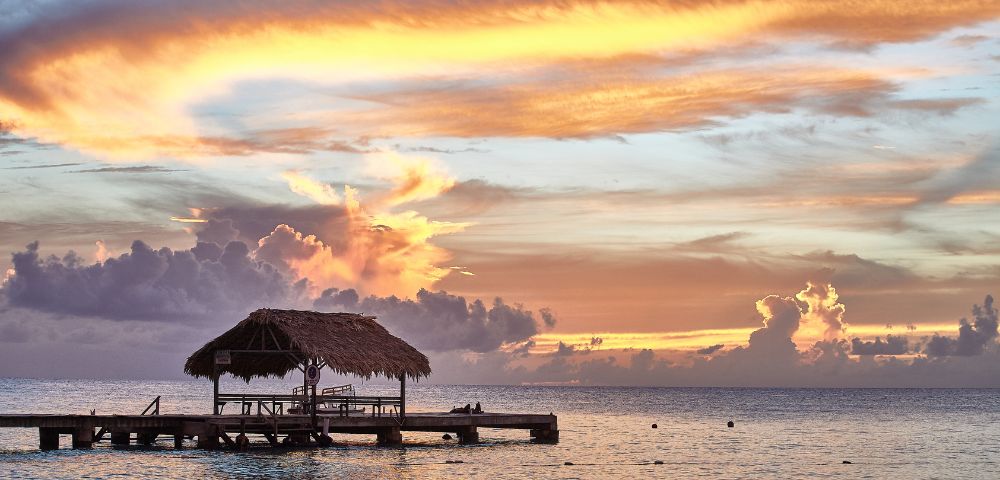 Tropical sunset over calm ocean, with a rustic wooden pier and thatched-roof hut in silhouette. The sky is vibrant with orange, pink, and blue hues.