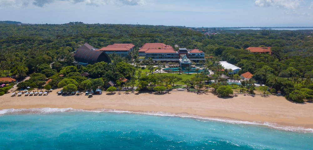 This image shows a stunning aerial view of a beachfront resort surrounded by lush greenery. At the bottom, there’s a long stretch of golden sandy beach with turquoise waves gently rolling onto the shore.