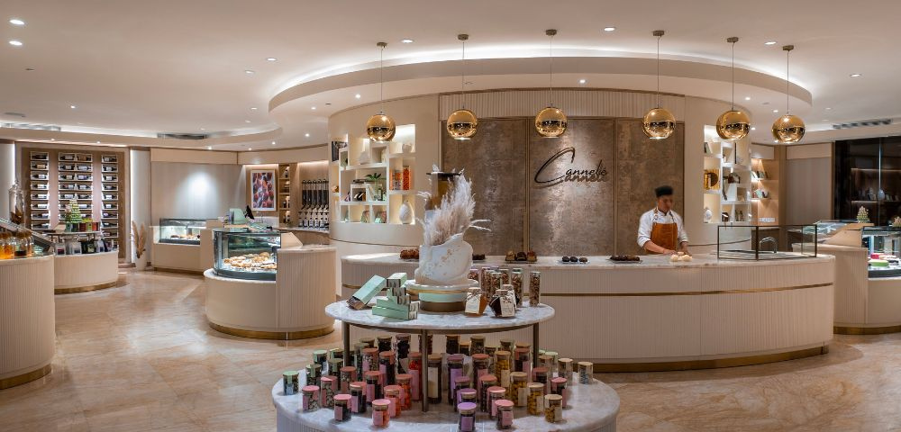 This image shows a luxurious and modern patisserie or dessert boutique. The space has a clean, elegant design with cream-colored walls, marble-style floors, and soft lighting that creates a warm and inviting atmosphere.
