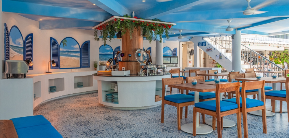 This image shows a bright and stylish restaurant interior with a Mediterranean-inspired design. The space is decorated in a blue-and-white color scheme, reminiscent of Greek island architecture.