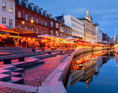 A vibrant evening scene along a canal with glowing red and orange lights reflecting in the water. People sit under umbrellas at outdoor cafes. Historic buildings line the street, creating a lively and inviting atmosphere.