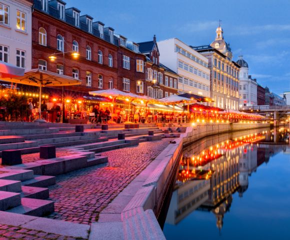 A vibrant evening scene along a canal with glowing red and orange lights reflecting in the water. People sit under umbrellas at outdoor cafes. Historic buildings line the street, creating a lively and inviting atmosphere.