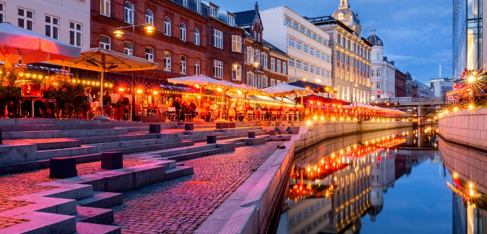 A vibrant evening scene along a canal with glowing red and orange lights reflecting in the water. People sit under umbrellas at outdoor cafes. Historic buildings line the street, creating a lively and inviting atmosphere.