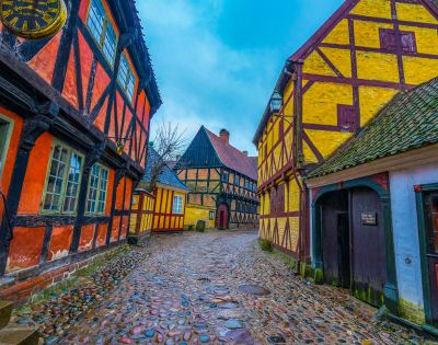 Cobblestone street lined with colorful, half-timbered houses in red, yellow, and black in an old European village under a cloudy blue sky.