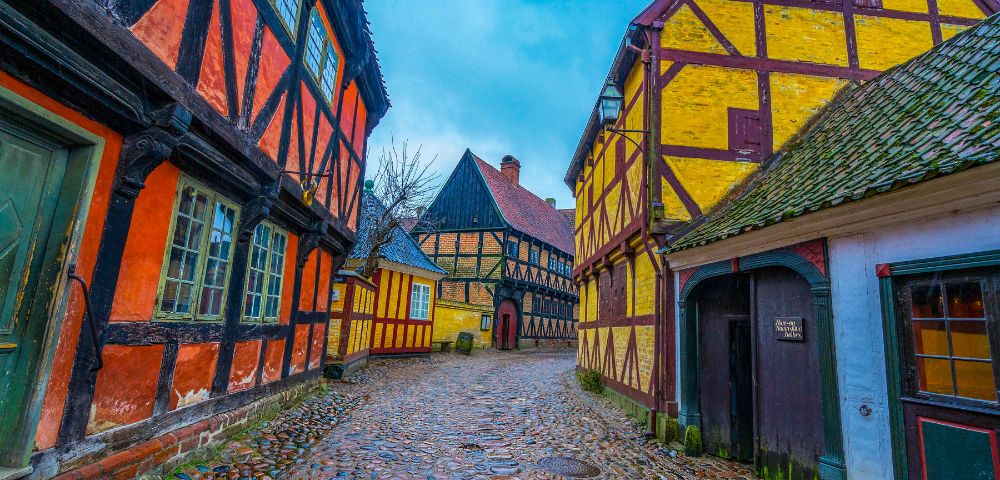 Cobblestone street lined with colorful, half-timbered houses in red, yellow, and black in an old European village under a cloudy blue sky.