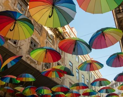 Colorful umbrellas suspended above a narrow street, creating a vibrant canopy. Surrounded by old buildings, it conveys a whimsical and lively atmosphere.