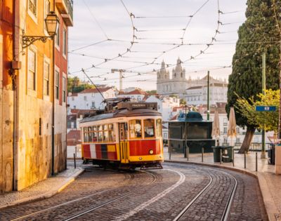 A vintage yellow and red tram on a cobblestone street in Lisbon, with warm sunlight illuminating nearby historic buildings. The scene feels nostalgic and picturesque.