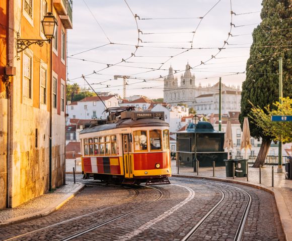 A vintage yellow and red tram on a cobblestone street in Lisbon, with warm sunlight illuminating nearby historic buildings. The scene feels nostalgic and picturesque.
