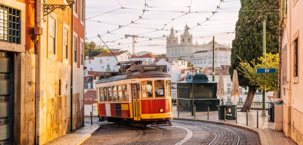 A vintage yellow and red tram on a cobblestone street in Lisbon, with warm sunlight illuminating nearby historic buildings. The scene feels nostalgic and picturesque.