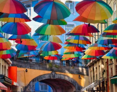 Numerous colorful umbrellas are suspended in the air, creating a vibrant canopy over a narrow street. The scene is lively and joyful.