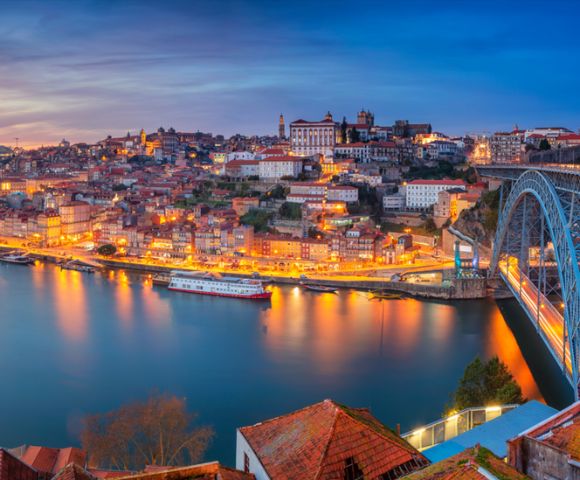 Scenic view of Porto, Portugal, during twilight. The city is illuminated with warm lights reflecting on the Douro River. A steel bridge is visible, enhancing the stunning panorama.
