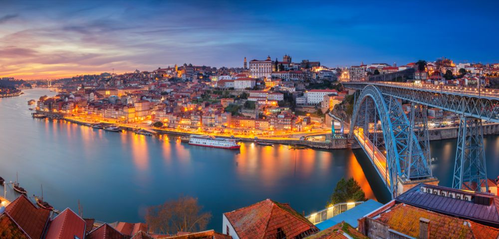Scenic view of Porto, Portugal, during twilight. The city is illuminated with warm lights reflecting on the Douro River. A steel bridge is visible, enhancing the stunning panorama.