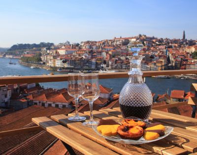 A wooden table set with a wine decanter, two glasses, and pastries overlooks a scenic riverside city under a clear blue sky, evoking a sense of relaxation.