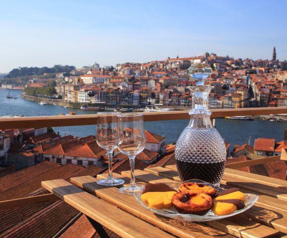 A wooden table set with a wine decanter, two glasses, and pastries overlooks a scenic riverside city under a clear blue sky, evoking a sense of relaxation.