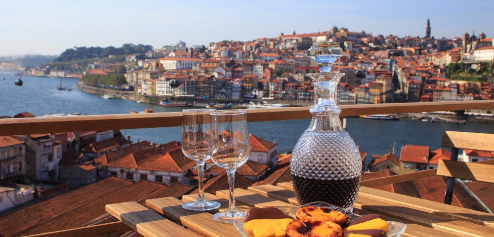 A wooden table set with a wine decanter, two glasses, and pastries overlooks a scenic riverside city under a clear blue sky, evoking a sense of relaxation.
