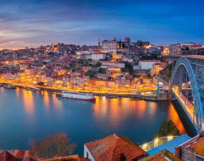 Scenic view of Porto, Portugal, during twilight. The city is illuminated with warm lights reflecting on the Douro River. A steel bridge is visible, enhancing the stunning panorama.