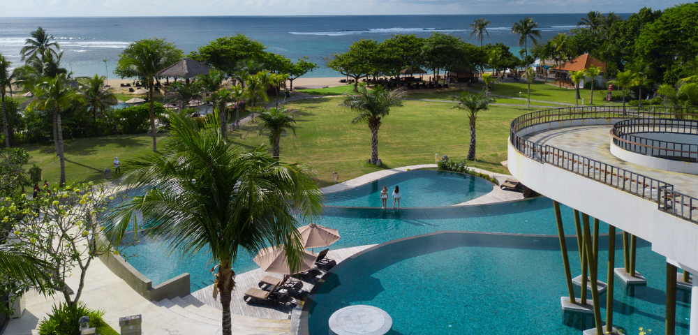 This image shows a beautiful tropical resort setting by the ocean. In the foreground, there’s a large, curvy infinity pool with different levels and shaded lounge chairs under umbrellas. Two people are standing in the shallow section of the pool, enjoying the view.