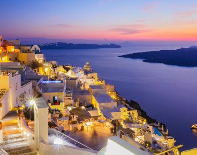 A stunning sunset view of a coastal town with white buildings and glowing lights overlooking a calm sea and distant islands.