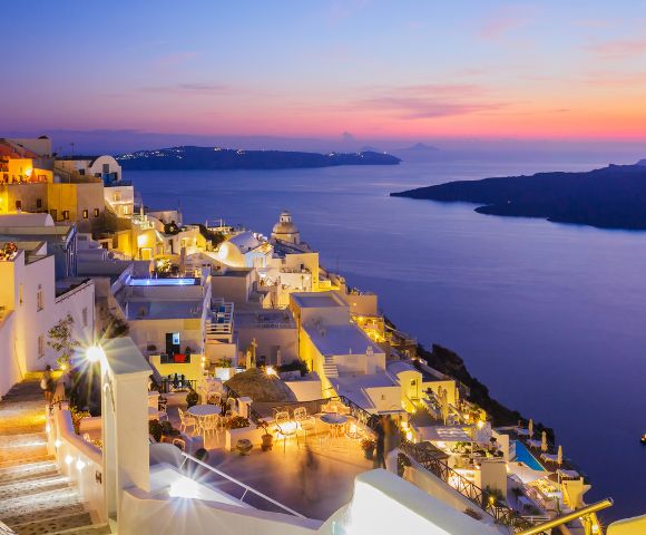 A stunning sunset view of a coastal town with white buildings and glowing lights overlooking a calm sea and distant islands.