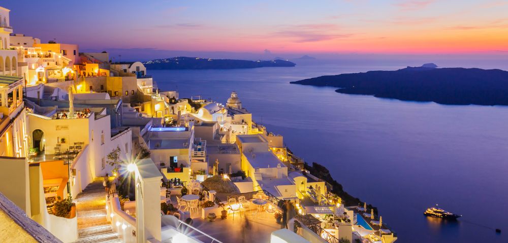 A stunning sunset view of a coastal town with white buildings and glowing lights overlooking a calm sea and distant islands.