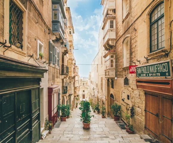 A charming narrow street in Valletta, Malta, featuring historic stone buildings, balconies, and potted plants under a bright blue sky.