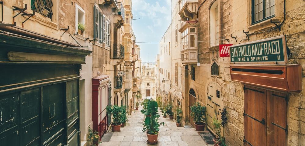 A charming narrow street in Valletta, Malta, featuring historic stone buildings, balconies, and potted plants under a bright blue sky.