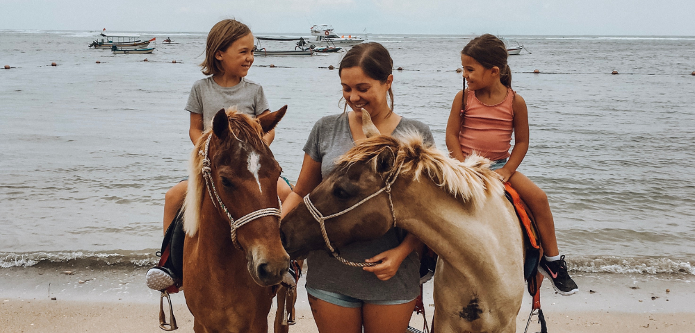 Two horses stand on a beach, with three children riding them. Fishing boats are visible in the background under a cloudy sky.