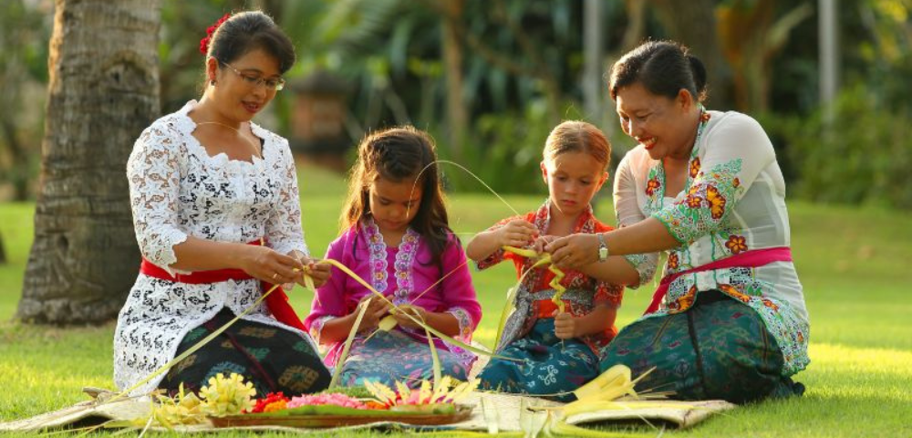 Four individuals are seated on grass, weaving traditional crafts with colorful materials and smiling, surrounded by lush greenery.