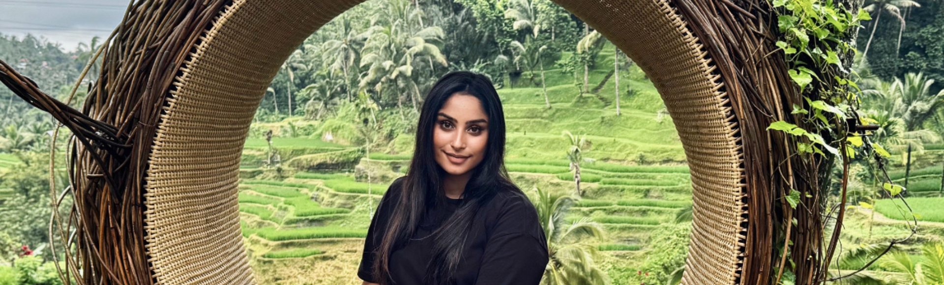 A person sits on a circular woven seat overlooking lush green rice terraces and palm trees in a scenic landscape.