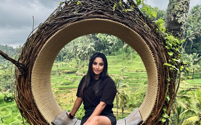 A person sits on a circular woven seat overlooking lush green rice terraces and palm trees in a scenic landscape.