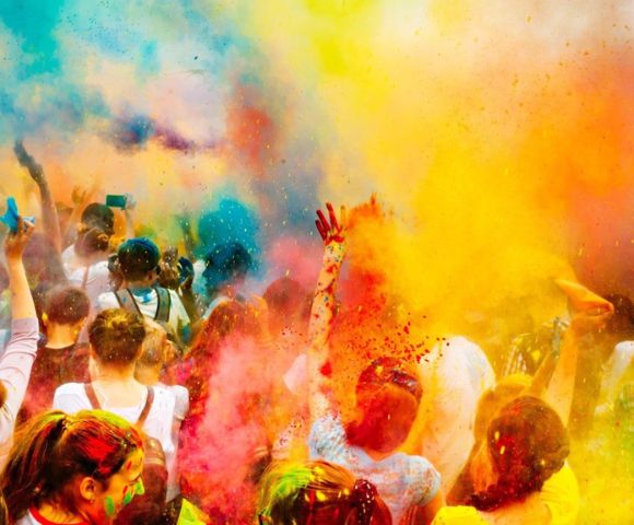 A vibrant scene of people celebrating a festival, throwing colorful powders into the air. Bright hues of yellow, orange, and pink create a joyful, energetic atmosphere.