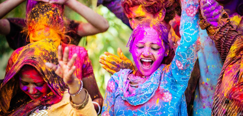 A joyful group celebrates Holi, with vibrant colors on their faces and clothes. Smiling people, lively gestures, and bright powders convey excitement.