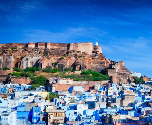 Fortress atop a hill overlooks a city of blue buildings under a vibrant blue sky. The scene conveys a historic and tranquil atmosphere.