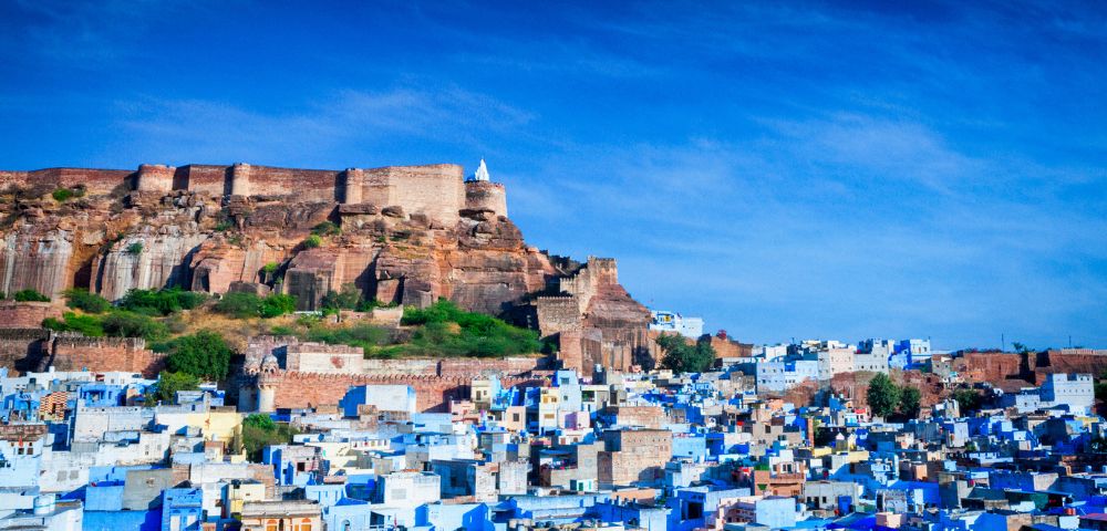 Fortress atop a hill overlooks a city of blue buildings under a vibrant blue sky. The scene conveys a historic and tranquil atmosphere.