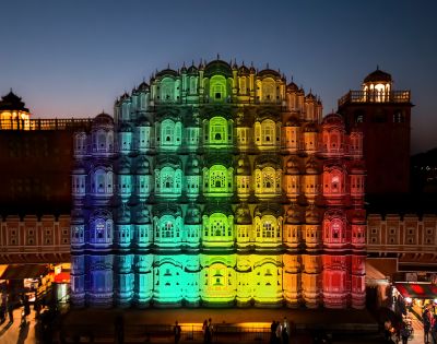Hawa Mahal illuminated in vibrant rainbow colors at dusk, with intricate windows, creating a festive, celebratory atmosphere. Silhouetted sky above.