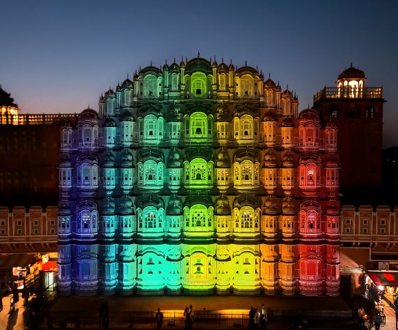 Hawa Mahal illuminated in vibrant rainbow colors at dusk, with intricate windows, creating a festive, celebratory atmosphere. Silhouetted sky above.